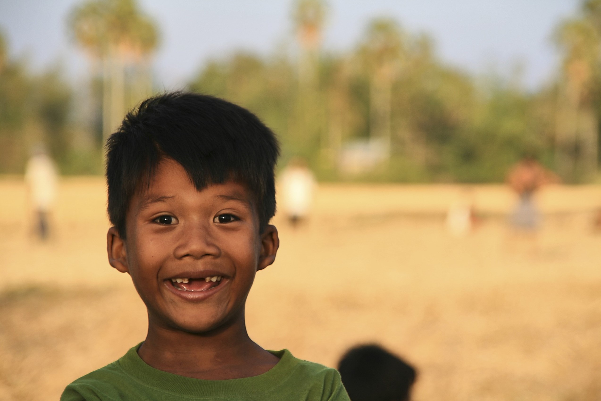 An image of a child standing in a landscape with trees and grass and people in the background which is all out of focus and so blurred. The child is boy about the age of 6 and is smiling and has two front teeth missing. 