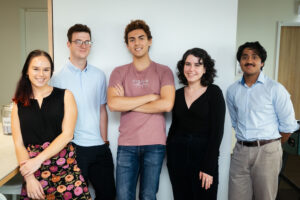 Two women and three men, all aged in their 20s, stand in front of a white wall.