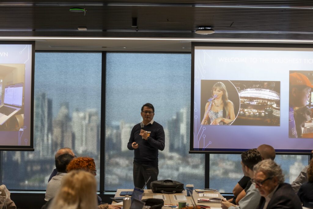 A man gives a presentation to a room. Behind him, a screen shows Taylor Swift. The skyline of Sydney is visible through the windows behind him.